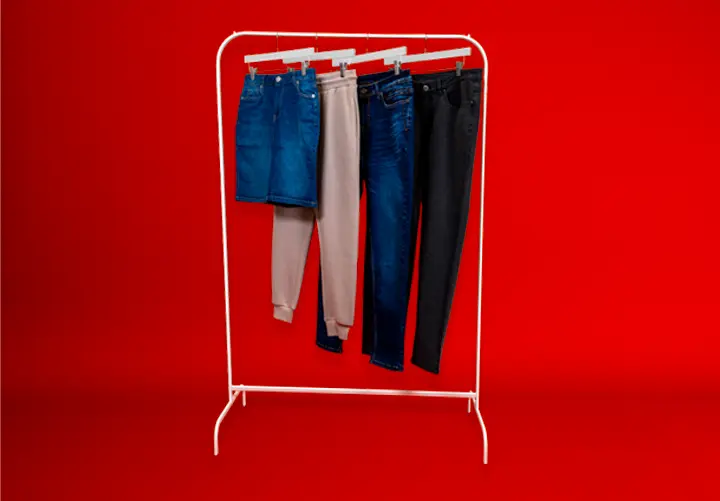 Ein Kleiderständer zeigt vier Hosen: hellblaue und schwarze Jeans, beige Jogginghosen und einen Jeansrock vor einem kräftig roten Hintergrund.